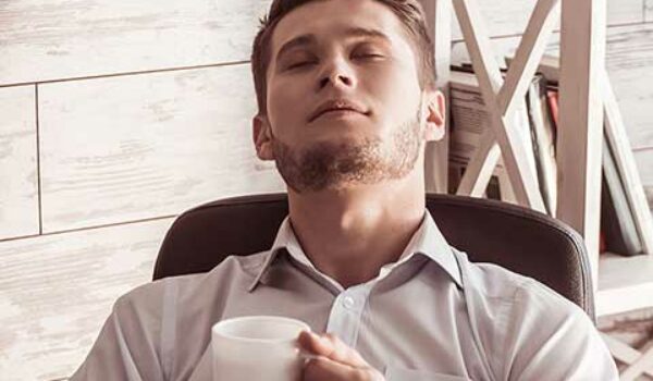 Mann sitzt mit einem Kaffee und geschlossenen Augen auf seinem Bürostuhl