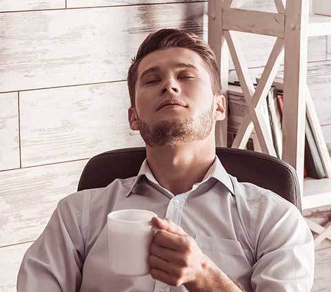 Mann sitzt mit einem Kaffee und geschlossenen Augen auf seinem Bürostuhl